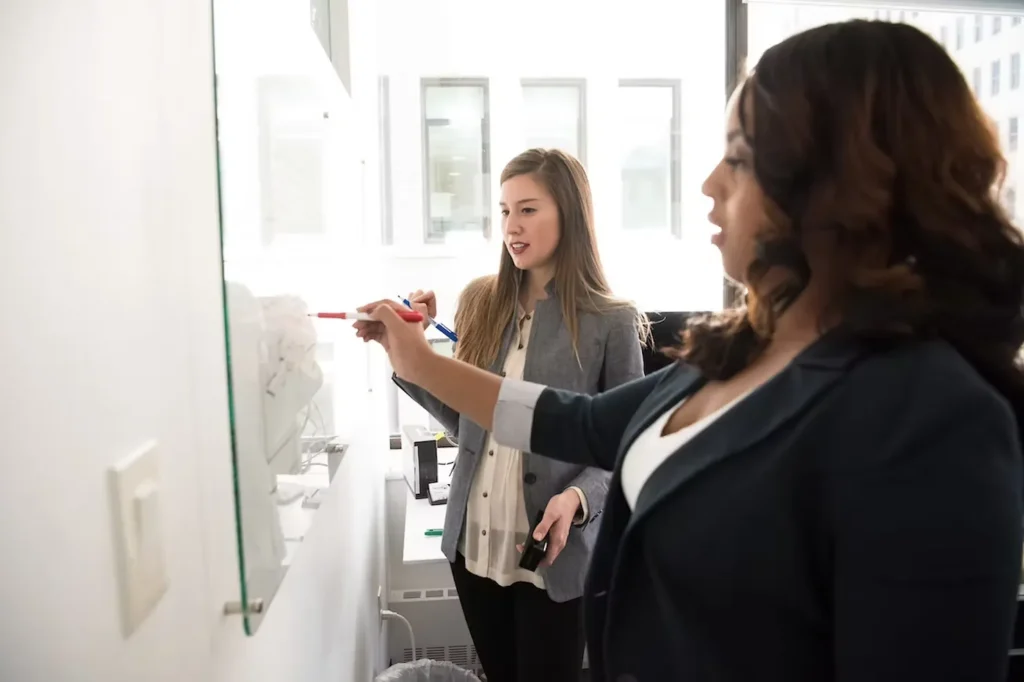 Two corporate women looking at a whiteboard with one of them writing something on the whiteboard